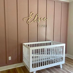 May include: A white crib with a wooden frame and a white mattress. The crib is in front of a pink wall with a gold wooden sign that says "Elsie Claire".