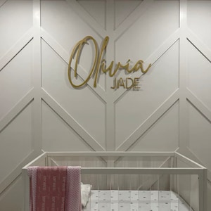May include: A white crib with a pink blanket and a gold name sign that reads "Olivia Jade" on a white wall with a geometric pattern.
