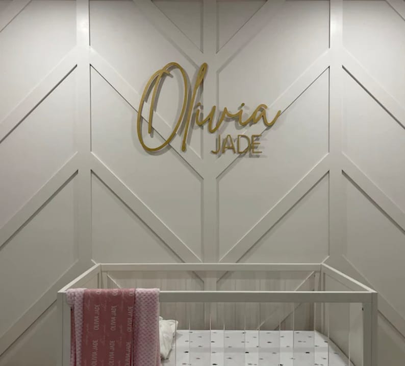 May include: A white crib with a pink blanket and a gold name sign that reads "Olivia Jade" on a white wall with a geometric pattern.
