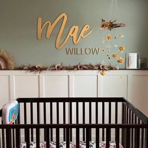 May include: A gold wooden name sign with the name "Mae Willow" above a crib with a floral mobile hanging above it.