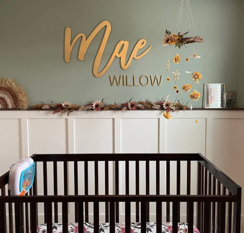May include: A gold wooden name sign with the name "Mae Willow" above a crib with a floral mobile hanging above it.
