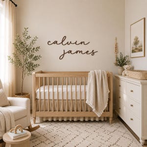 May include: A wooden crib with a white mattress and a beige tasseled blanket, positioned below a dark brown wall-mounted sign featuring the name calvin james in a cursive script font, set within a room containing a wooden rocking chair and dresser.