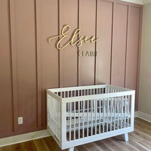 May include: A white crib with a wooden frame and a white mattress. The crib is in front of a pink wall with a wooden name sign that says "Elsie Claire".