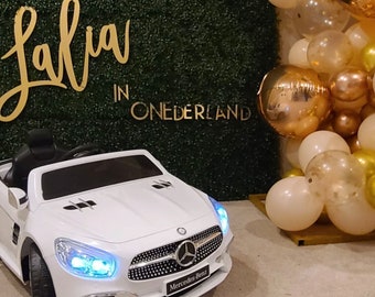 May include: A white Mercedes-Benz toy car with blue headlights is positioned in front of a green hedge wall. Gold lettering on the wall reads "Lalia in ONEDERLAND". Balloons in gold, white, and clear colors are to the right.