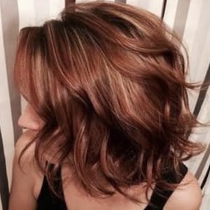 Puede incluir: Primer plano de cabello en capas, hasta los hombros, con una base marrón rojizo y reflejos rubios más claros. El cabello está peinado con ondas suaves y raya a un lado, creando un aspecto voluminoso. La persona lleva una camiseta negra.