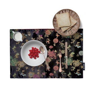 May include: Black placemat with a gold and colourful floral pattern. A white plate with red raspberries, a white creamer, a fork, a knife, and a spoon are on the placemat. The placemat has a red border and the text "Made in North" is printed on the bottom right corner.