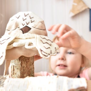 May include: A child holds up a handmade owl toy with brown eyes and wings. The owl is made of beige fabric and has a brown felt beak. The owl is sitting on a wooden log.