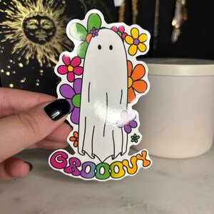 Può includere: Un fantasma bianco con un testo "Groovy" arcobaleno e una corona di fiori con fiori rosa, viola, arancioni e gialli.