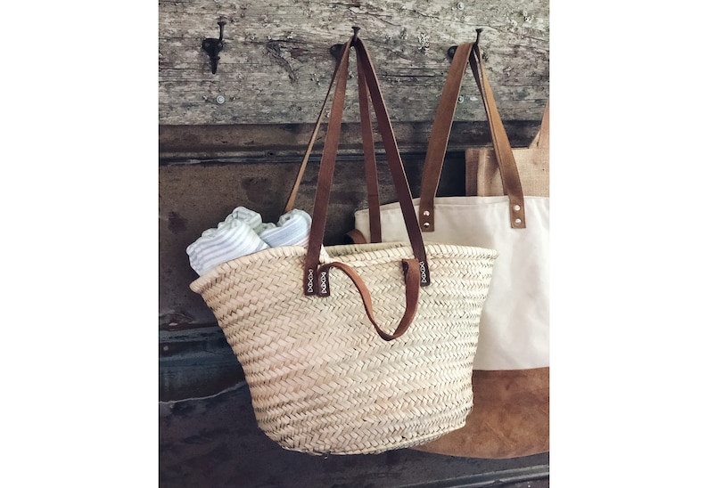 Op de afbeelding: Een geweven strooien tas met bruine leren handvatten en het woord "boogie" gedrukt op de handvatten. De tas hangt aan een houten muur met een witte canvas tas ernaast.