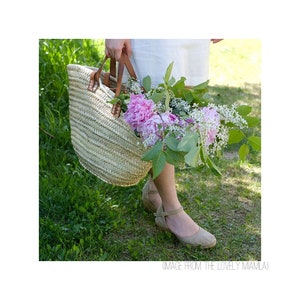 Op de afbeelding: Een vrouw draagt een geweven mand gevuld met roze pioenen en witte bloemen. Ze draagt een witte jurk en beige sandalen.