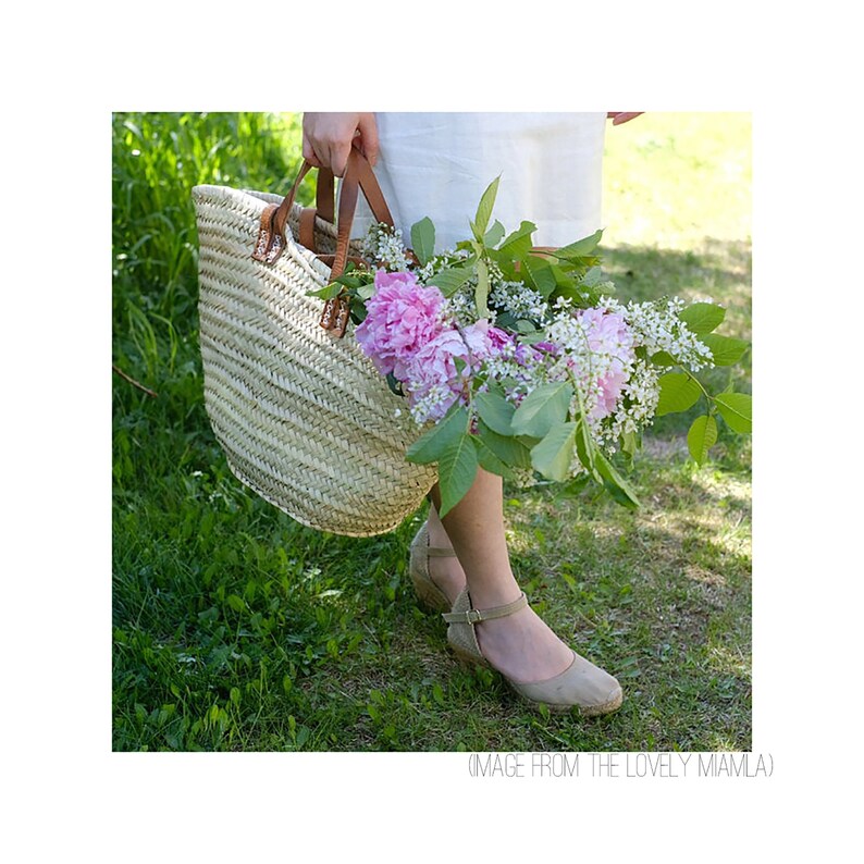 Op de afbeelding: Een vrouw draagt een geweven mand gevuld met roze pioenen en witte bloemen. Ze draagt een witte jurk en beige sandalen.