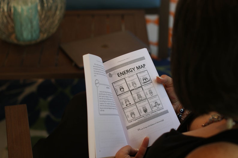 May include: A woman is reading a book titled "Energy Map" with a grid of nine squares, each containing a cartoon owl with a different expression. The book is open to a page with the text "Your Organizational Clarity Guiding Compass".