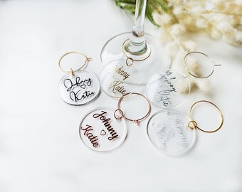 May include: Six clear acrylic wine glass charms with gold wire rings. Each charm has a different name written in black script, including "Johnny & Katie".