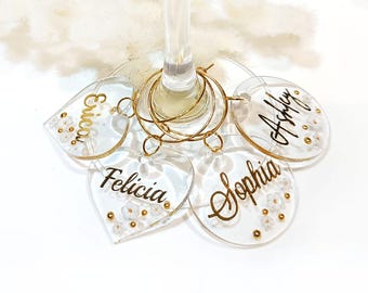 May include: Four clear acrylic wine glass charms with gold lettering and white beads. The charms are labelled "Erica", "Felicia", "Sophia", and "Ashley".