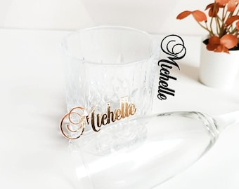 May include: Two clear glass cups with the name "Michelle" written on them in gold and black lettering. The cups are on a white surface.