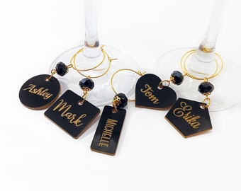 May include: Set of five black acrylic wine glass charms with gold lettering. The charms are shaped as a circle, square, rectangle, heart, and a hexagon. The names Ashley, Mark, Michelle, Tom, and Erika are written on the charms.