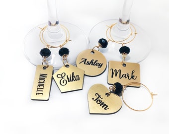 May include: Set of five gold-coloured wine glass charms with black beads and personalised names: Michelle, Erika, Ashley, Mark, and Tom.
