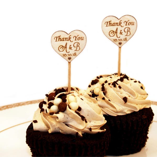 May include: Two chocolate cupcakes with brown frosting and chocolate sprinkles topped with wooden heart-shaped signs that say "Thank You A & B 10.10.18".