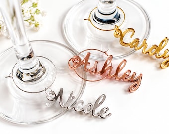 May include: Three metal wine glass charms in gold, rose gold, and silver tones. Each charm features a circular wire loop and a dangling script nameplate reading Carolyn, Ashley, or Nicole, attached to the base of clear glass stems.