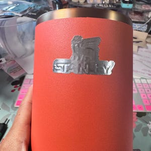 May include: A coral-colored stainless steel tumbler with a silver logo that reads "STANLEY". The tumbler has a textured finish and a silver rim. The image is a close-up of the tumbler.