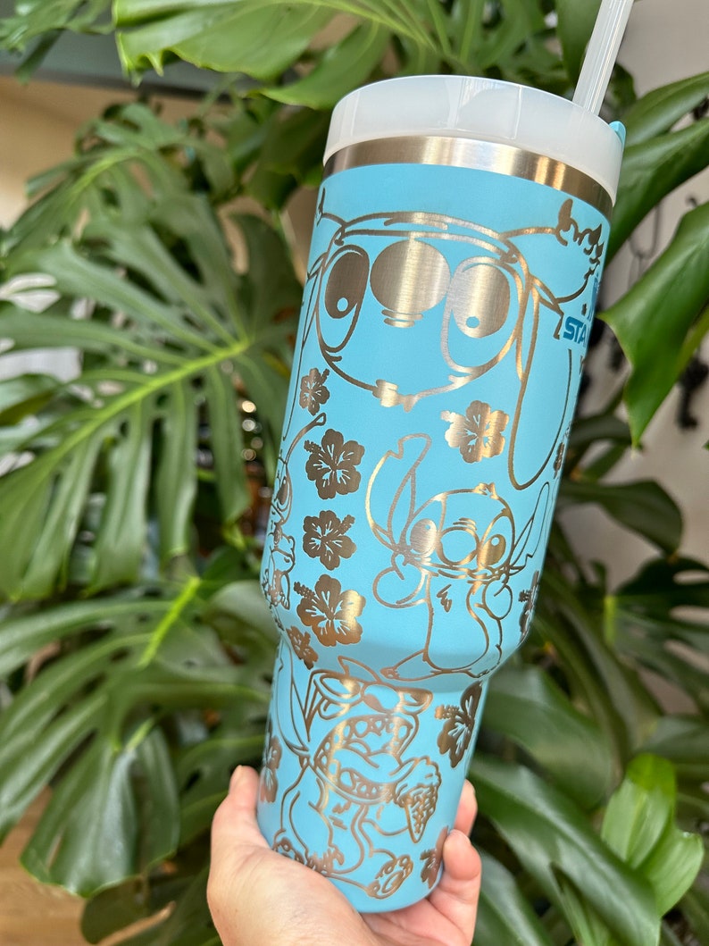 May include: A light blue stainless steel tumbler with a gold metallic design featuring Stitch and other characters from the Disney movie Lilo & Stitch. The tumbler has a straw and a lid.
