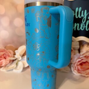 May include: A bright blue insulated tumbler with a large side handle, a clear lid, and a straw. The exterior features laser-etched line art of Lilo and Stitch characters surrounded by tropical flowers and leaves, with the Stanley logo visible.