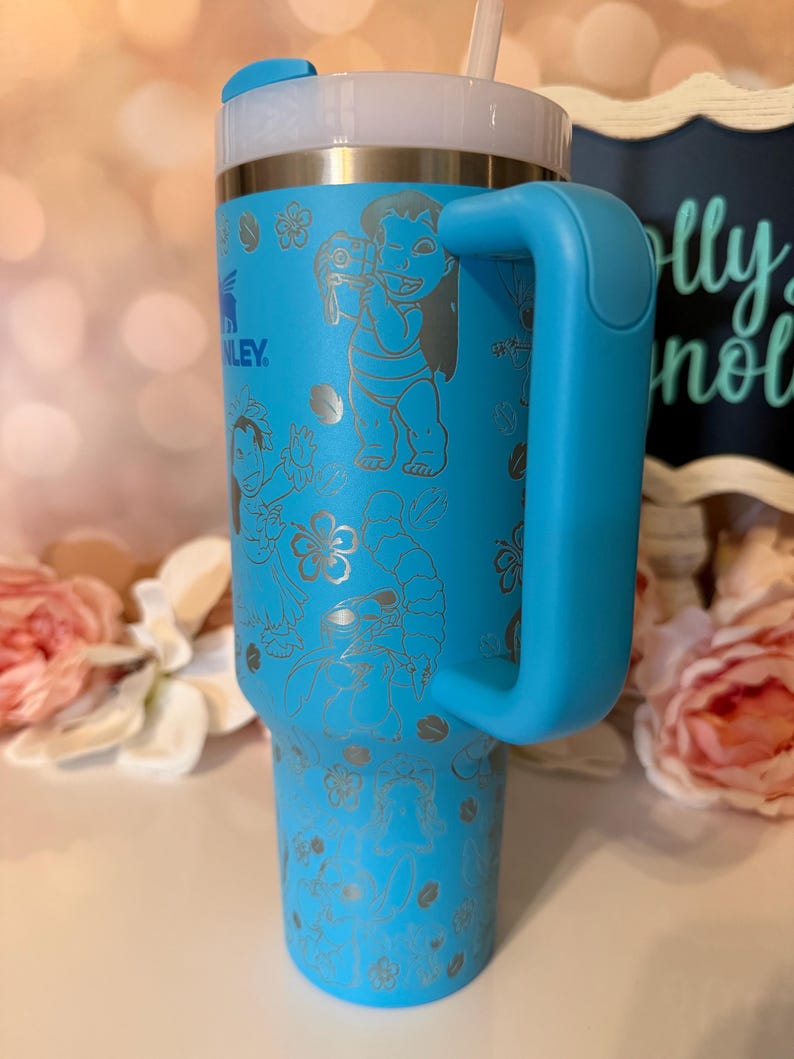 May include: A bright blue insulated tumbler with a large side handle, a clear lid, and a straw. The exterior features laser-etched line art of Lilo and Stitch characters surrounded by tropical flowers and leaves, with the Stanley logo visible.