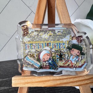 Puede incluir: Un amuleto de acr&iacute;lico transparente con una ilustraci&oacute;n de dibujos animados de una casa amarilla con dos figuras delante. Las figuras llevan ropa de invierno y una de ellas sostiene un bast&oacute;n de caramelo rojo y blanco. El amuleto est&aacute; en un caballete de madera.