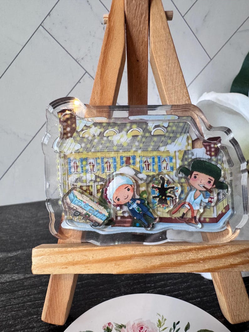 Puede incluir: Un amuleto de acr&iacute;lico transparente con una ilustraci&oacute;n de dibujos animados de una casa amarilla con dos figuras delante. Las figuras llevan ropa de invierno y una de ellas sostiene un bast&oacute;n de caramelo rojo y blanco. El amuleto est&aacute; en un caballete de madera.
