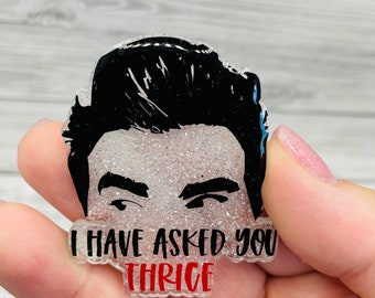 Könnte beinhalten: Ein durchsichtiger Acryl-Anstecker mit Glitzer-Finish, der eine schwarze Grafik von Haaren und Augenbrauen zeigt. Der Text "I HAVE ASKED YOU THRICE" ist in schwarzer und roter Schrift auf dem unteren Teil des Designs aufgedruckt.