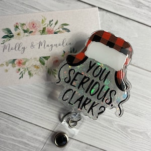 May include: A clear plastic badge holder with a red and black checked design. The holder features a white background with the text "You Serious Clark?" in black lettering. The holder is attached to a clear plastic retractable badge reel.