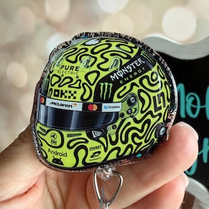 May include: A neon yellow and black racing helmet badge reel featuring a complex abstract pattern. The helmet displays logos for Monster Energy, McLaren, OKX, Android, and Pure Electric, with a dark visor and a metallic clip attachment at the base.