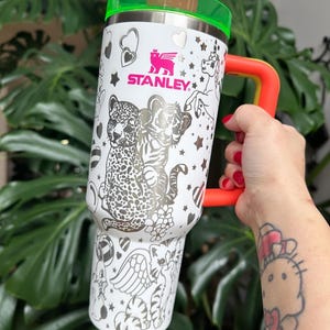 Puede incluir: Un vaso Stanley blanco con tapa verde ne&oacute;n y asa de color naranja brillante. El exterior presenta ilustraciones de l&iacute;neas negras de un leopardo, un tigre, un unicornio y varios corazones y estrellas. Una pajita rosa est&aacute; insertada en la tapa.