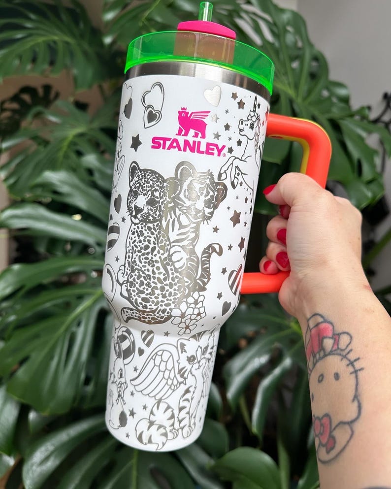 Puede incluir: Un vaso Stanley blanco con tapa verde ne&oacute;n y asa de color naranja brillante. El exterior presenta ilustraciones de l&iacute;neas negras de un leopardo, un tigre, un unicornio y varios corazones y estrellas. Una pajita rosa est&aacute; insertada en la tapa.
