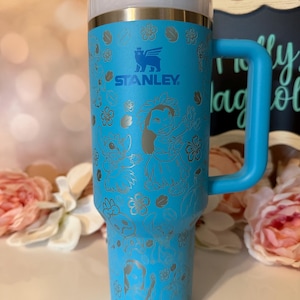 May include: A bright blue Stanley tumbler with a handle, featuring a laser-etched pattern of Lilo and Stitch characters surrounded by tropical flowers. The cup has a clear lid with a blue flip-top closure and a straw, displaying the Stanley logo.