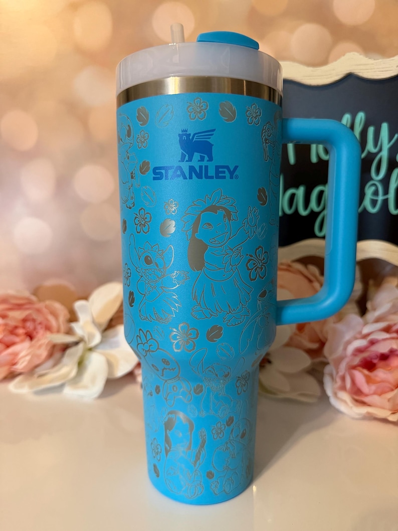 May include: A bright blue Stanley tumbler with a handle, featuring a laser-etched pattern of Lilo and Stitch characters surrounded by tropical flowers. The cup has a clear lid with a blue flip-top closure and a straw, displaying the Stanley logo.