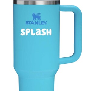 May include: A bright turquoise Stanley tumbler with a large side handle, a white lid, and a clear straw. The word "SPLASH" is printed in white letters on the front, positioned below the blue Stanley logo featuring a winged bear emblem.