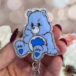 May include: A light blue Care Bear badge reel featuring Grumpy Bear. The badge reel has a clear plastic backing and a retractable cord. The Grumpy Bear design has a white belly with a cloud, and a glittery outline.
