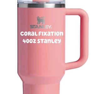 Puede incluir: Un vaso Stanley de color coral de 40oz con asa, tapa transparente y pajita. La taza presenta el logotipo de Stanley y el texto 'CORAL FIXATION 40oz STANLEY' impreso en blanco en la parte frontal. El dise&ntilde;o incluye una base c&oacute;nica y un asa lateral.
