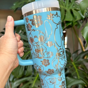 May include: A light blue stainless steel tumbler with a handle and a straw lid. The tumbler features a gold metallic design of Stitch and other characters from the Disney movie Lilo & Stitch. The design also includes gold metallic hibiscus flowers.