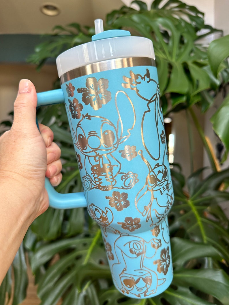 May include: A light blue stainless steel tumbler with a handle and a straw lid. The tumbler features a gold metallic design of Stitch and other characters from the Disney movie Lilo & Stitch. The design also includes gold metallic hibiscus flowers.