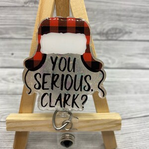 May include: A clear plastic badge holder with a red and black plaid Christmas hat design and the text "You serious, Clark?"  The badge holder has a black retractable reel.