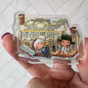 May include: A clear plastic ornament featuring a cartoon illustration of a yellow house with blue shutters, a snow-covered roof, and two figures standing in front of it. The figures are wearing winter clothing and holding gifts. The ornament is decorated with glitter and snowflakes.