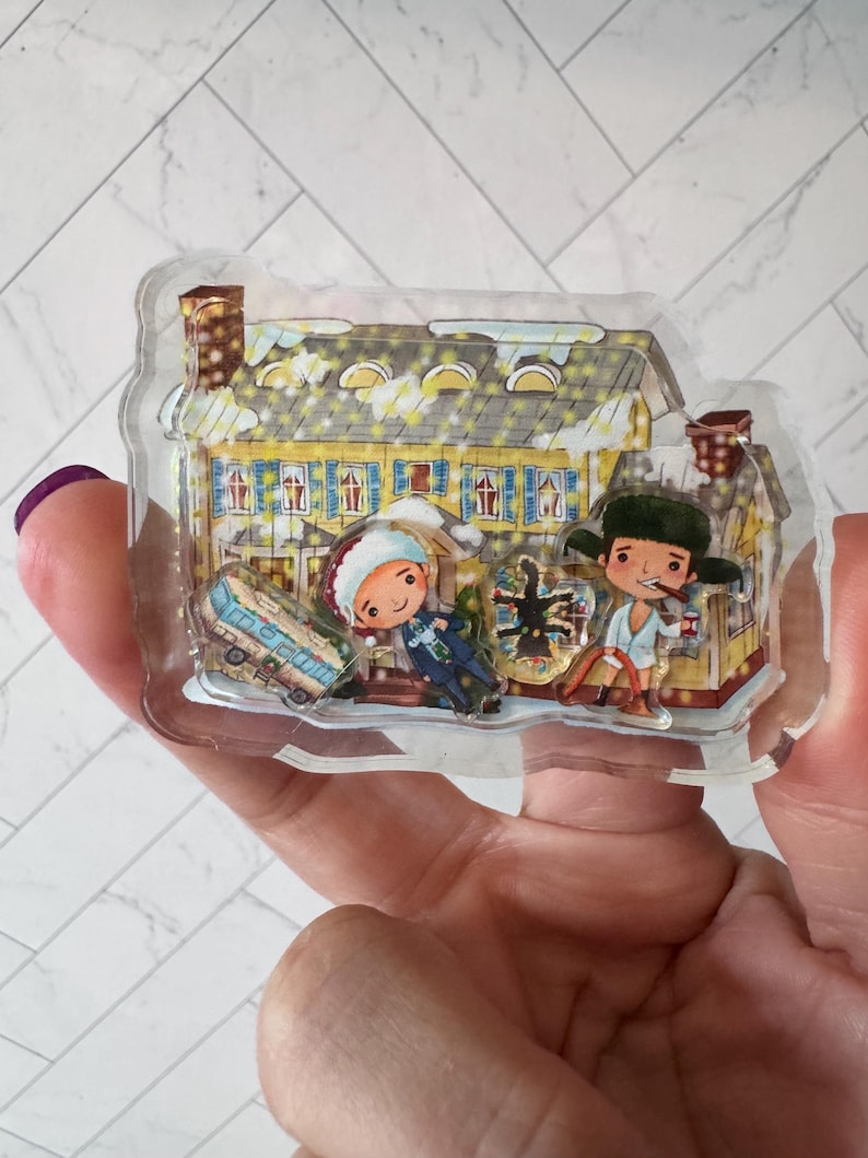 Puede incluir: Un adorno de pl&aacute;stico transparente con una ilustraci&oacute;n de dibujos animados de una casa amarilla con contraventanas azules, un techo cubierto de nieve y dos figuras de pie frente a ella. Las figuras llevan ropa de invierno y sostienen regalos. El adorno est&aacute; decorado con purpurina y copos de nieve.