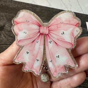 May include: A pink glitter resin badge holder with a large pink bow and white ghost silhouettes. The bow has a white lace trim and the badge holder has a clear plastic clip.