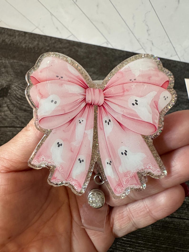 May include: A pink glitter resin badge holder with a large pink bow and white ghost silhouettes. The bow has a white lace trim and the badge holder has a clear plastic clip.