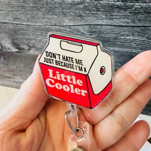 May include: A clear plastic badge holder with a red and white design. The design features a cartoon image of a milk carton with the text "Don't hate me just because I'm a Little Cooler".