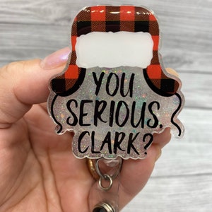 May include: A red and black checked Christmas hat with a white brim and the text "You Serious Clark?" on a sparkly silver background. The hat is attached to a clear plastic badge holder with a black swivel clip.