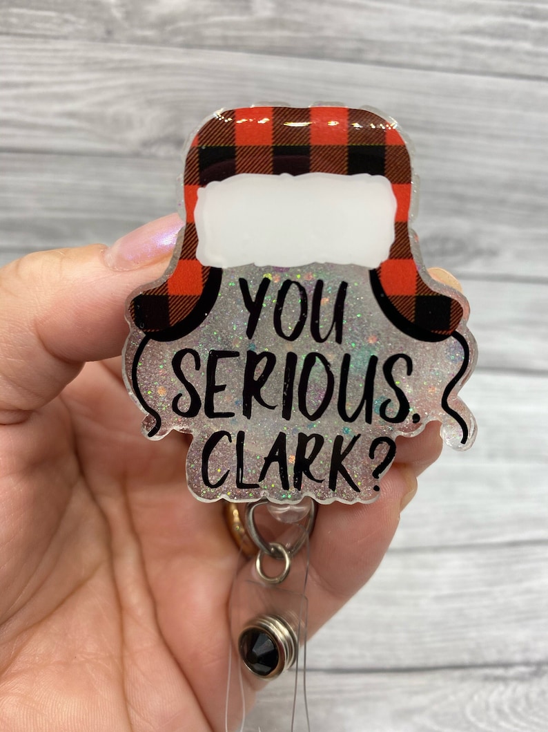 May include: A red and black checked Christmas hat with a white brim and the text "You Serious Clark?" on a sparkly silver background. The hat is attached to a clear plastic badge holder with a black swivel clip.