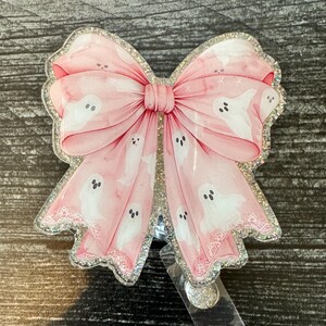 May include: A pink glitter badge holder with a large pink bow and white ghost silhouettes. The bow has a white lace trim.